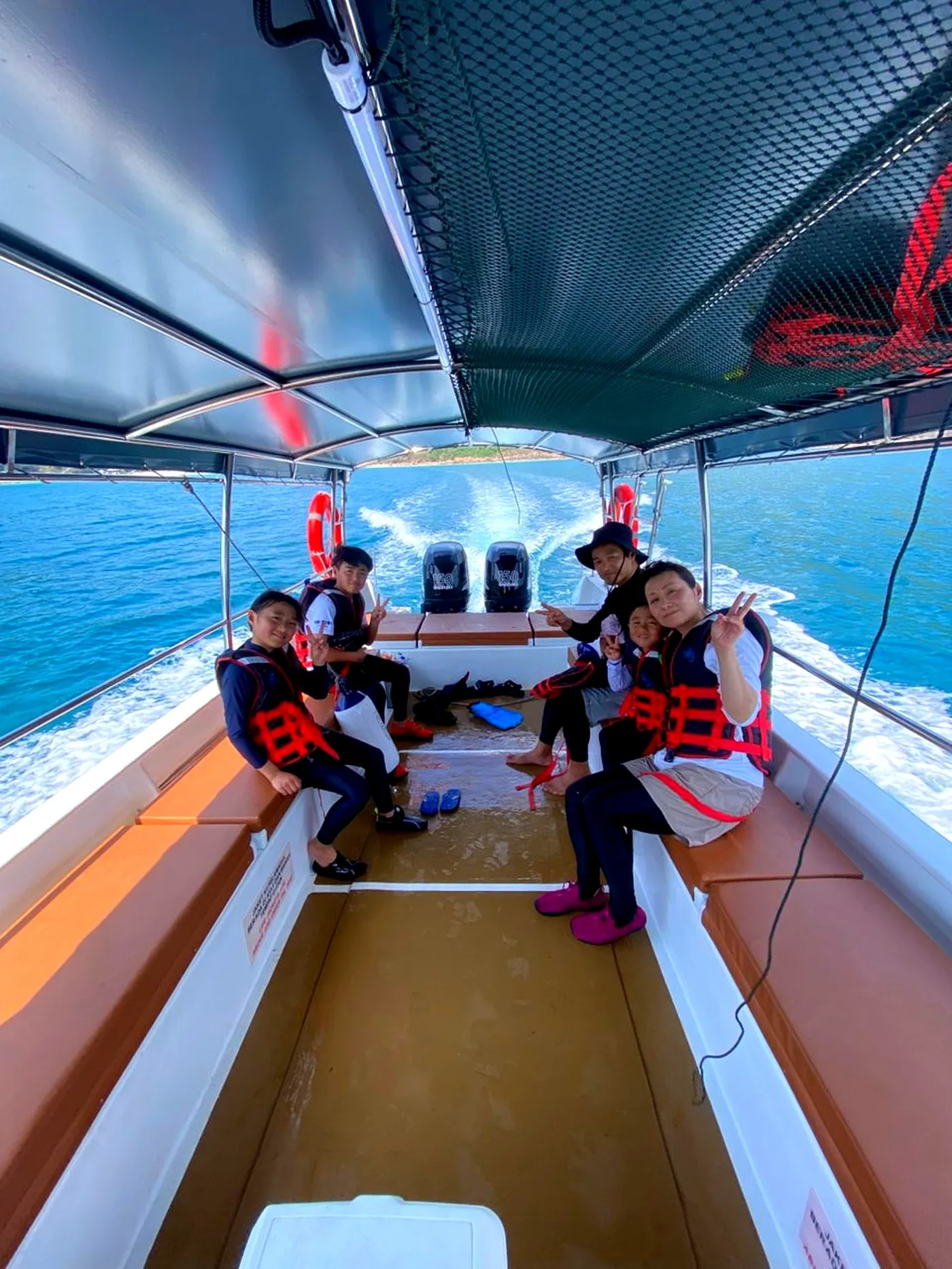A family with young kids in life jackets smiling and giving peace signs at the back of the boat, sea wake trailing behind