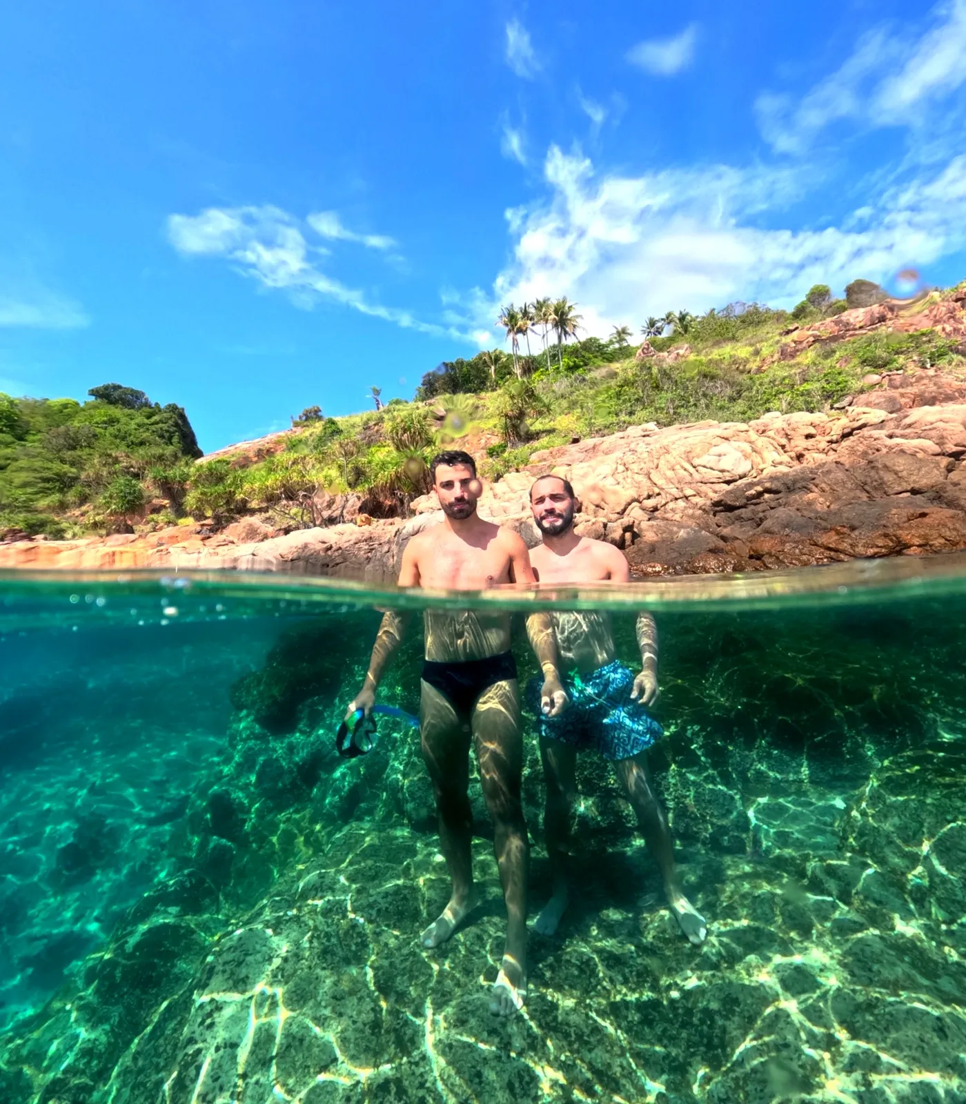 Half-underwater split shot of two snorkelers with a rocky shoreline and shimmering sand below