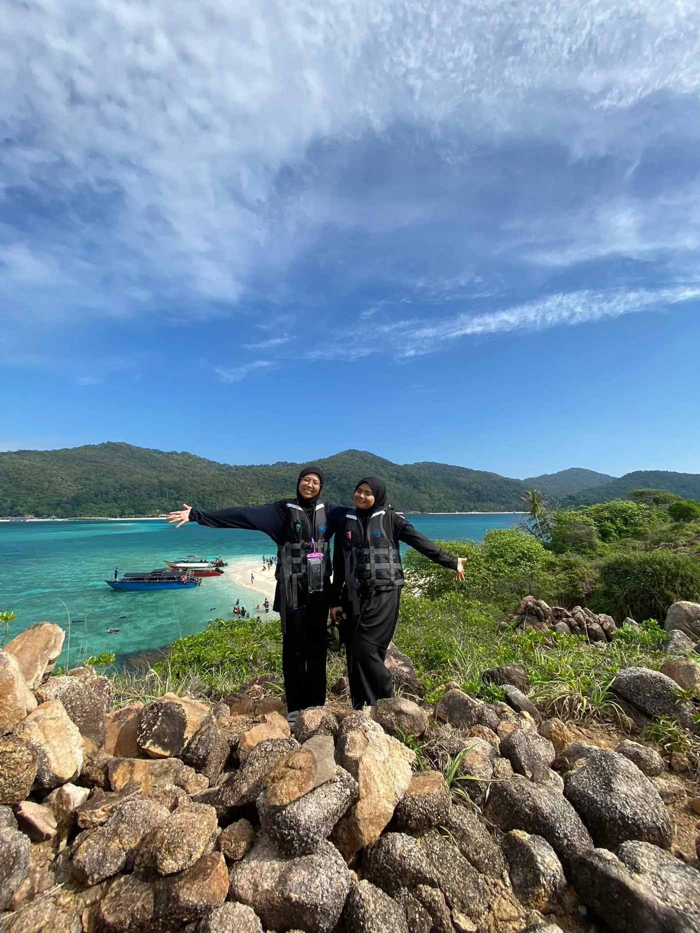 Two guests smiling from a rocky overlook with the boat, sandbank, and turquoise lagoon below